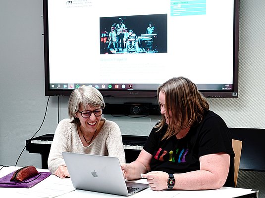 Zwei Personen sitzen nebeinander an einem Tisch, darauf sind Unterlagen und ein Laptop